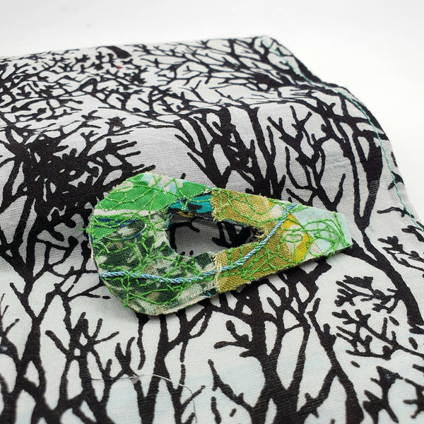 An 80's design fabric brooch made with a mixture of different shades of greens resting on a white and black piece of fabric 