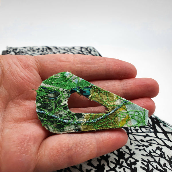 An 80's design fabric brooch made with a mixture of different shades of greens being held in a white woman's hand