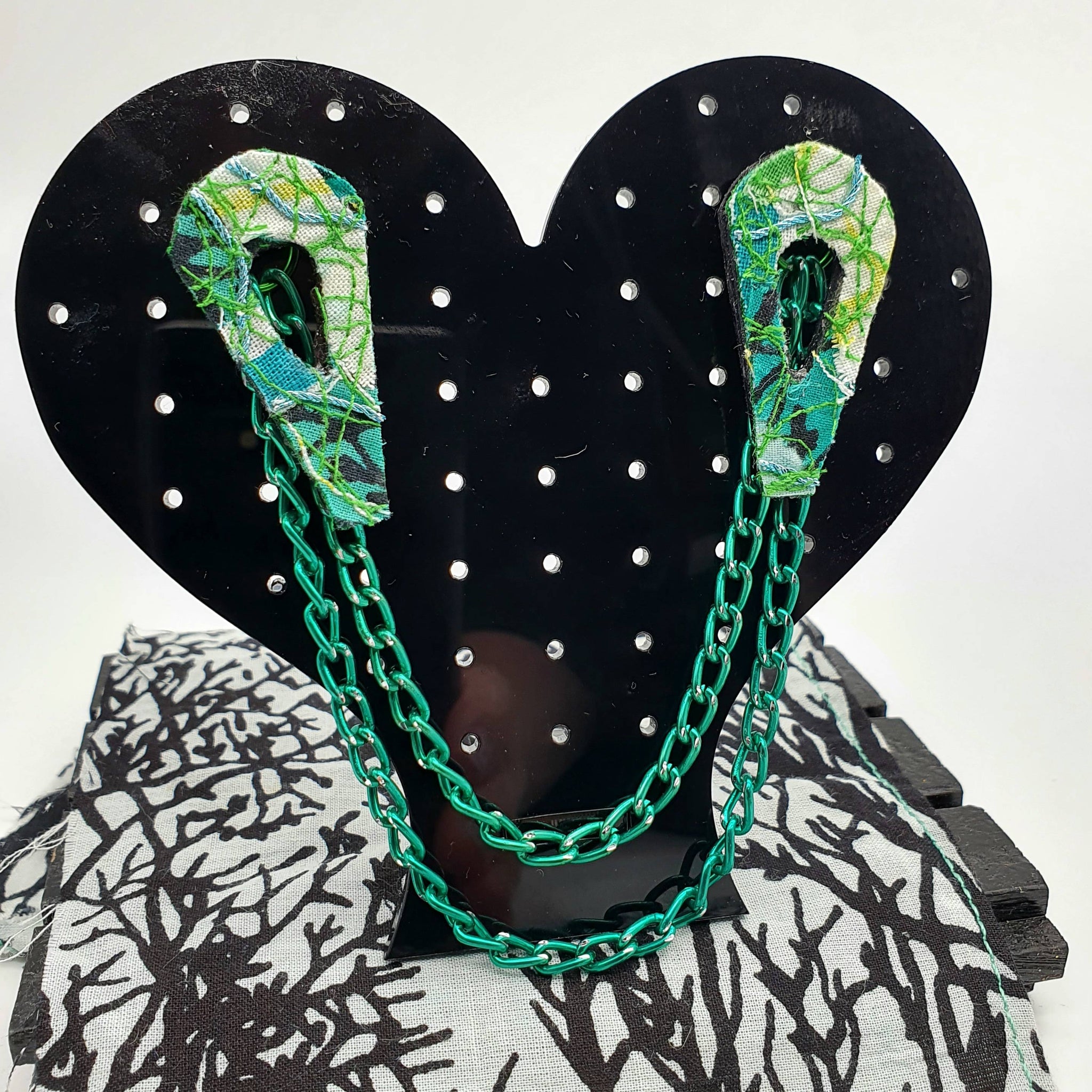 An 80's design fabric collar pin set made with a mixture of different shades of greens resting on a black acrylic heart shaped display stand.