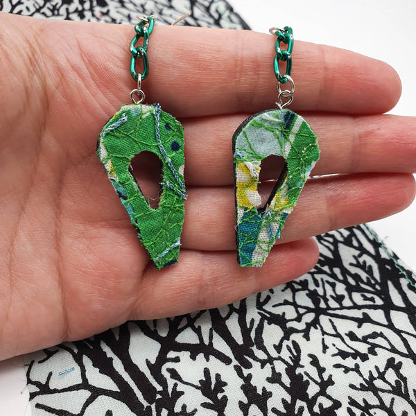 A pair of dangle style earrings hanging on a green chain held in a white woman's hand.