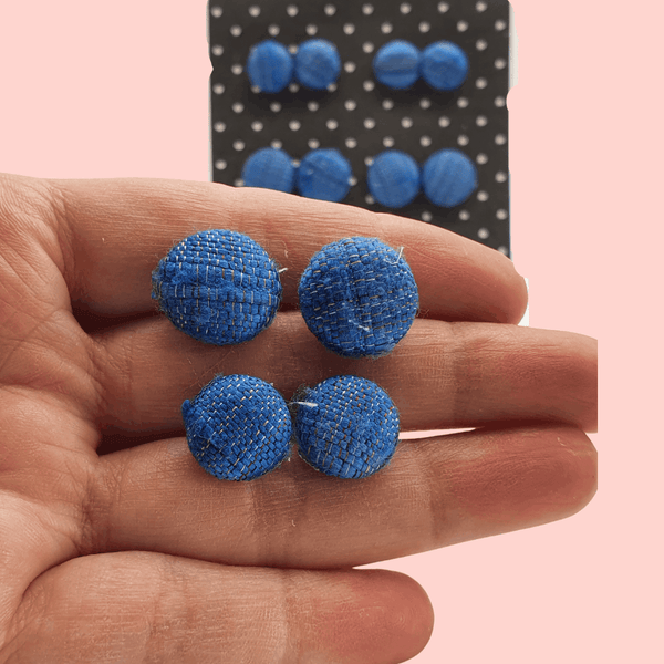 A white woman's hand holding two pairs of blue fabric button stud earrings.