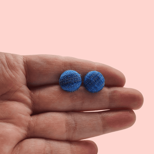 A white woman's hand holding a pair of blue fabric button stud earrings.