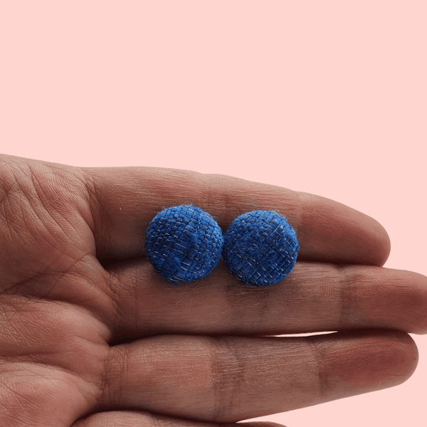 A white woman's hand holding a pair of blue fabric button stud earrings.